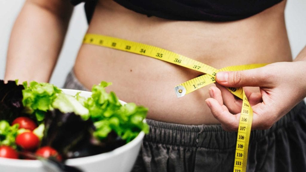 Person measuring waist with a yellow tape measure, holding a fresh salad bowl, focusing on weight loss