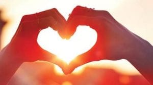 Hands forming a heart shape in front of the sun during sunset