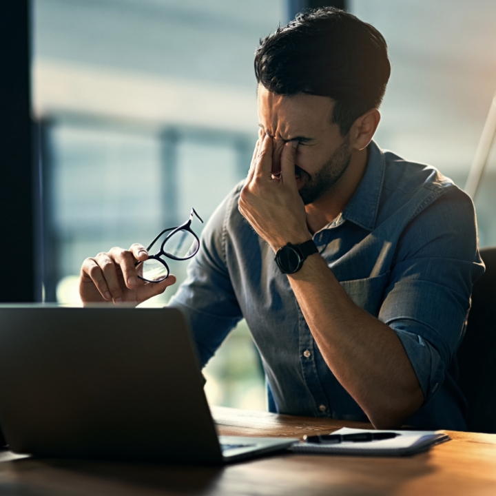 Man working at a desk with a laptop, holding eyeglasses and appearing thoughtful