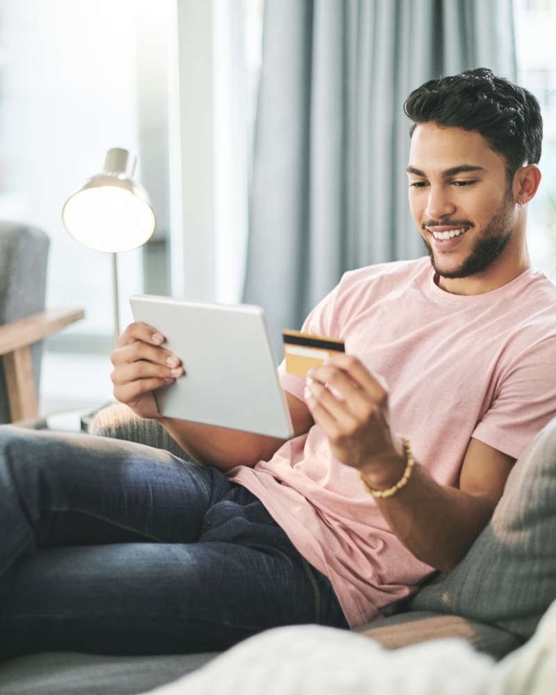 A man relaxes on a sofa, using a tablet and holding a credit card, representing the Pricing and Value for Money