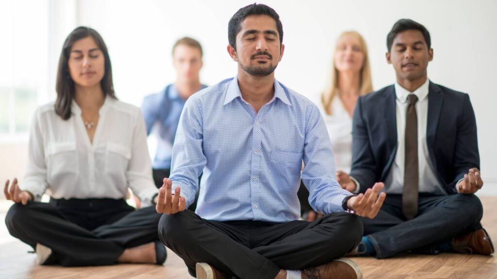 Group of business professionals sitting cross-legged on the floor in a bright office, practicing meditation together as part of a corporate wellness program.