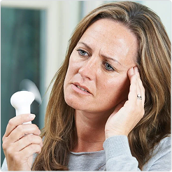 Woman holding a handheld fan to her face, appearing to cool down, representing menopause symptoms.
